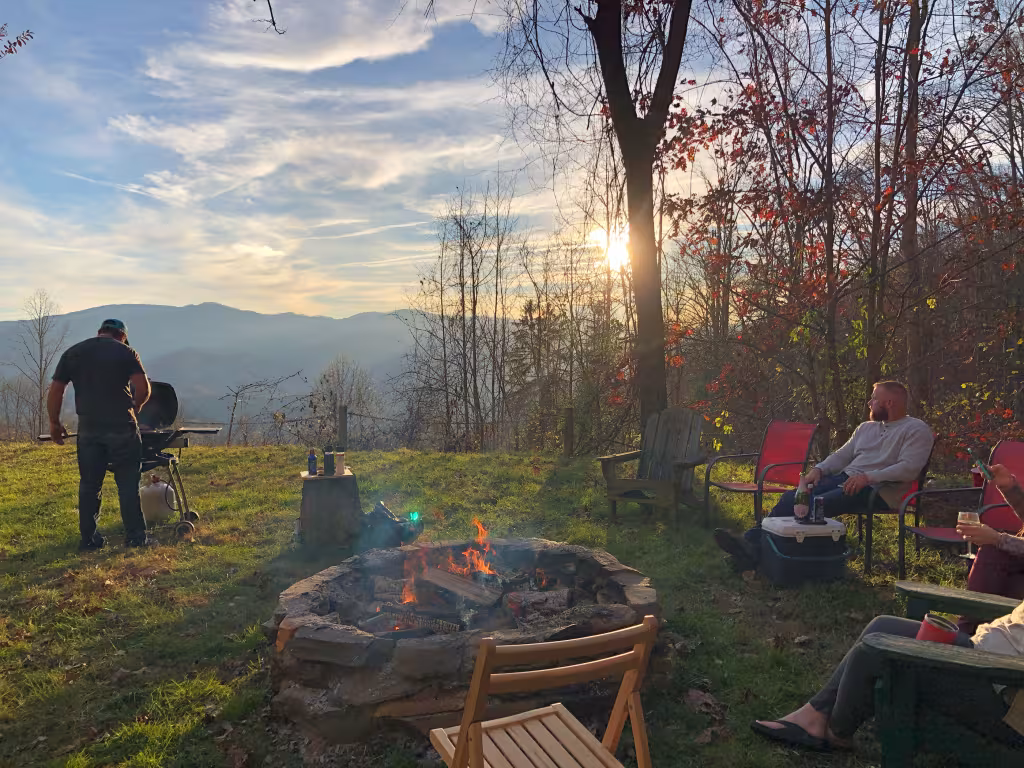 Guests around the stone fire pit on the lawn at sunset, one at the grill, with the mountains and fall color behind them